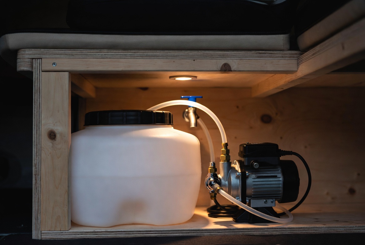 Depósito de agua y bomba 12V instalados bajo un banco de madera en el interior de una furgoneta